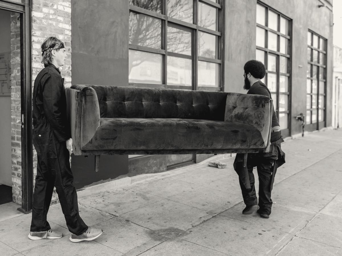 Two crew members in dark work clothes carrying a sofa out of a building